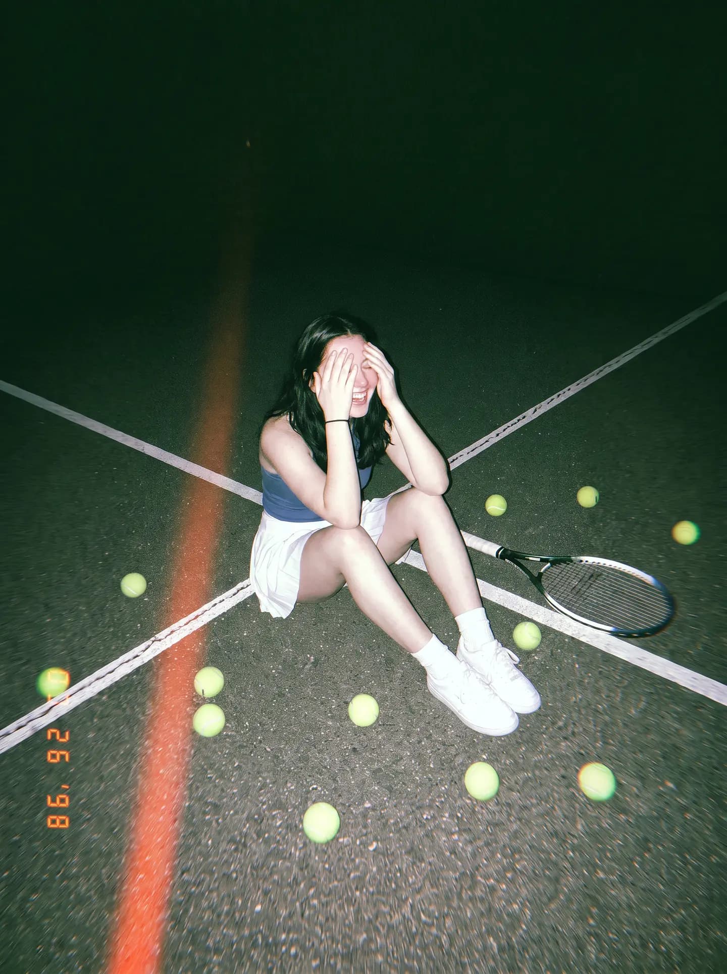 A tennis player sitting on a floodlit court at night, laughing, balls scattered around her